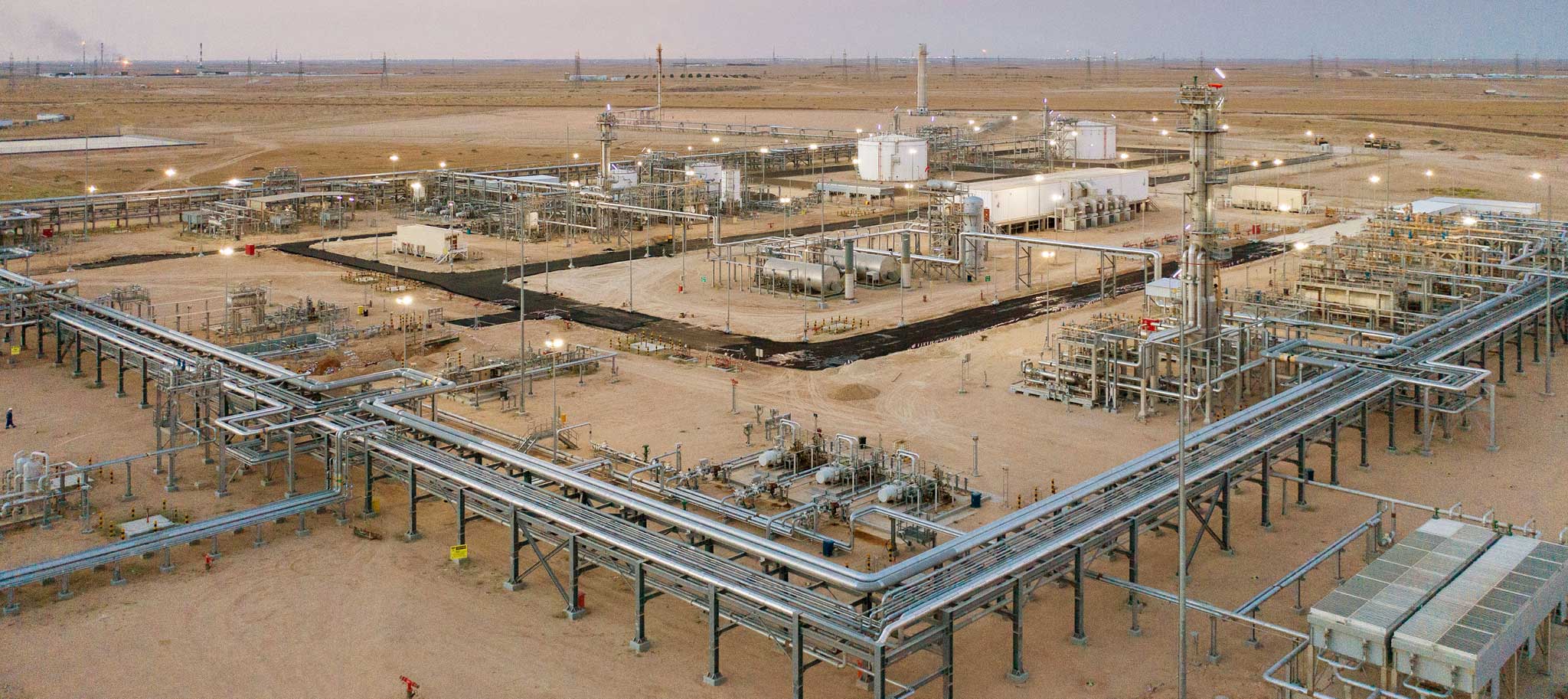 Aerial view of a large oil and gas processing facility in a desert landscape, with interconnected pipelines, storage tanks, and industrial structures under a colorful evening sky.